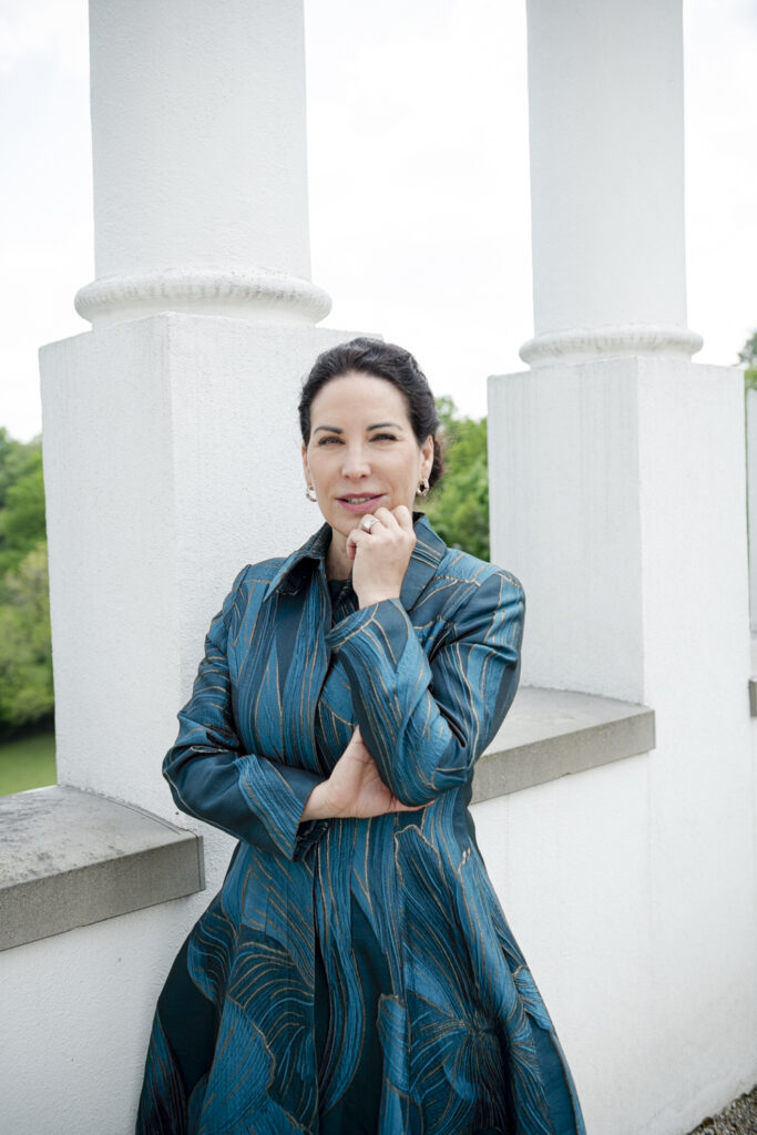 Eine dunkelhaarige Frau im türkisen Mantel (Prof. Dr. Katrin Winkler) im Vöhlinschloss in Illertissen vor einer weißen Säule.