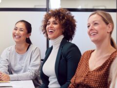 Drei Frauen mit unterschiedlichen Herkünften sitzen in einem Büro und lachen.