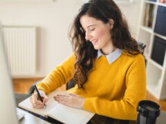 Eine Frau mit braunen Locken und einem gelben Pullover arbeitet von zu Hause aus und macht Notizen