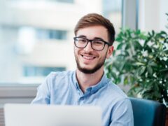 Ein junger Mann mit Brille arbeitet an seinem Laptop und lächelt dabei.