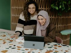 Zwei Frauen an einem Computer, die eine sitzt am Tisch vor einem Microsoft Surface, sie trägt ein Schaila und lacht, schräg hinter ihr steht die zweite Frau, sie trägt eine Brille und hat braune Locken, sie lacht ebenfalls