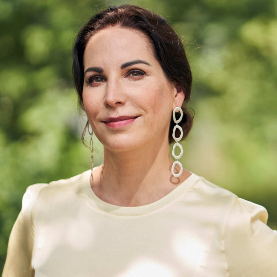 Portrait von Prof Dr Katrin Winkler, lange Statement-Ohrringe, helles T-Shirt, natürliches Licht, grüner Hintergrund
