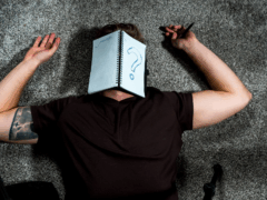 Eine männliche Person liegt auf dem Boden, auf seinem Gesicht ein aufgeschlagenes Notizbuch mit einem großen Fragezeichen. Er weiß nicht, wie wissenschaftlich arbeiten funktioniert