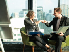 Zwei Personen sitzen an einem Tisch mit Computern auf dem Schoß, sie fist bumpen, ein Symbol für Jobsharing in der Führung