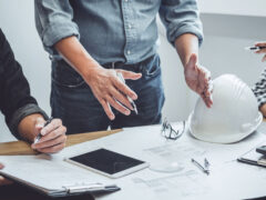 Man sieht die Hände von drei Personen, einen Helm und einen Tisch mit Unterlagen. praxisnahe technikweiterbildung für betriebswirte