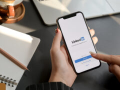 Ein Bild, um LinkedIn Profil optimieren darzustellen. Zu sehen ist eine Hand, die ein Handy hält, auf dem Display ist die Startseite der LinkedIn-App. Im Hintergrund ein Notizbuch.