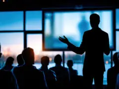 Das Bild zeigt eine professionelle Vortrags- oder Konferenzsituation in einer modernen, abendlichen Atmosphäre. Das Foto ist von hinten aus den Zuschauerreihen aufgenommen und arbeitet stark mit Silhouetten und Kontrasten. Im rechten Vordergrund steht die dunkle Silhouette eines Redners im Anzug, der dem Publikum zugewandt ist. Er hat den linken Arm leicht angehoben und die Hand geöffnet, um das Gesagte mit einer erklärenden Geste zu untermalen. Vor ihm sitzt im Halbdunkel das Publikum – eine Gruppe von mehreren Personen, die alle als dunkle Silhouetten von hinten zu sehen sind und aufmerksam nach vorne blicken. Der Hintergrund wird von einer riesigen, bodentiefen Fensterfront dominiert, durch die man im weichen, bläulichen Abendlicht vage eine Skyline oder Uferpromenade mit fernen Lichtern erahnen kann. Mittig im Hintergrund hängt eine große Präsentationsleinwand, die hell erleuchtet ist und ein unscharfes Bild zeigt. Durch das Zusammenspiel aus dem kühlen, blauen Licht von draußen und den warmen Lichtpunkten der Stadt wirkt die Szene sehr fokussiert, hochprofessionell und stimmungsvoll.