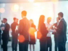 Das Bild zeigt eine stark verschwommene Networking-Szene im Querformat. Zu sehen sind Silhouetten zahlreicher Personen in eleganter Business-Kleidung, die in kleineren Gruppen beieinanderstehen und sich unterhalten – vermutlich auf einem Event, einer Konferenz oder einem Empfang. Das Bild ist in kühlen Blau- und Grautönen gehalten, wird aber im Zentrum durch warme, gelb-orange Lichteinfälle und glänzende Lichtpunkte durchbrochen. Die bewusste Unschärfe erzeugt eine elegante, professionelle und zeitlose Atmosphäre, die ideal für Themen rund um Networking, Karriere oder Unternehmensevents steht.