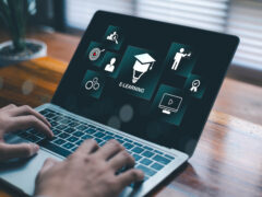 Das Bild zeigt eine Person, die an einem Laptop auf einem Holzschreibtisch arbeitet, wobei im Vordergrund die Hände beim Tippen zu sehen sind. Der Bildschirm präsentiert eine Reihe digitaler, blau-grün leuchtender Kacheln zum Thema Online-Bildung. Im Zentrum steht ein großes Symbol, das eine Glühbirne mit einem Doktorhut kombiniert, direkt darunter steht das Wort „E-LEARNING“. Drumherum gruppieren sich kleinere Icons wie eine Zielscheibe, Zahnräder, ein Präsentations-Symbol, ein Video-Player und ein Zertifikat. Die Szene wirkt modern und fokussiert.