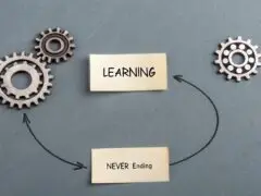 Das Bild zeigt eine symbolische Darstellung zum Thema lebenslanges Lernen auf einer grauen, strukturierten Oberfläche. In der Mitte liegen zwei gelbe Zettel übereinander: Der obere ist mit „LEARNING“ beschriftet, der untere mit „NEVER Ending“. Zwei feine, gezeichnete Pfeile verbinden die Elemente zu einem angedeuteten Kreislauf. Links und rechts befinden sich metallische Zahnräder – zwei ineinandergreifende Zahnräder auf der linken Seite und ein einzelnes Rad auf der rechten Seite. Das Motiv symbolisiert auf einfache Weise, dass Lernen ein kontinuierlicher, nie endender Prozess ist.