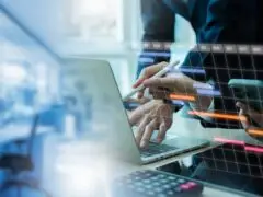 Das Bild zeigt eine moderne Arbeitsnetzwerk-Szene im Breitbildformat, die Themen wie Projektmanagement, Zeitplanung und Digitalisierung aufgreift. Im Vordergrund tippt eine Person an einem Laptop und zeigt mit einem Eingabestift auf den Bildschirm, während eine zweite Person daneben ein Smartphone hält. Über die Szene gelegt ist ein virtuelles Gantt-Diagramm mit Monatsleisten und farbigen Projektbalken. Links verschmilzt das Bild sanft mit einer hellen, leicht verschwommenen Büroansicht in kühlen Blautönen. Das Motiv vermittelt Effizienz, strukturierte Planung und moderne Zusammenarbeit.
