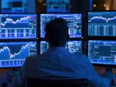 Das Bild zeigt eine Person von hinten, die in einem abgedunkelten Raum vor einer Wand aus sechs großen Monitoren sitzt. Auf den Bildschirmen sind komplex strukturierte Finanzcharts, Aktienkurse und Datenanalysen in leuchtend blauen und grünen Tönen zu sehen. Die Szene ist komplett in kühles, blaues Monitorlicht getaucht und vermittelt eine hochkonzentrierte, moderne Arbeitsatmosphäre rund um Themen wie Trading, Datenanalyse, Finanzmärkte oder IT-Monitoring.