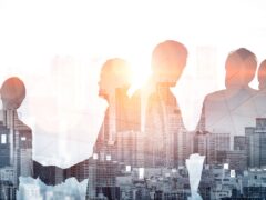 Das Bild zeigt eine Doppelbelichtung im breiten Panoramaformat zum Thema Business und Partnerschaft. Im Hintergrund steht die weitläufige, helle Silhouette einer Großstadt mit Wolkenkratzern. Im Vordergrund sind die schattigen Konturen mehrerer Personen in Business-Kleidung zu sehen, die direkt mit den Hochhäusern der Stadt verschmelzen. In der linken Hälfte schütteln sich zwei der Personen die Hände, was Verträge, Zusammenarbeit oder Fusionen symbolisiert. Das Bild wird von sanftem, warmem Sonnenlicht durchflutet und wirkt sehr professionell, global und zukunftsorientiert.