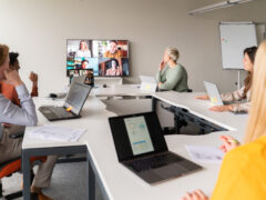 Das Bild zeigt ein hybrides Meeting in einem hellen, modernen Konferenzraum. Eine Gruppe von Teilnehmenden sitzt an U-förmig angeordneten, weißen Tischen mit geöffneten Laptops und Unterlagen, während an der Stirnseite des Raumes ein großer Monitor installiert ist. Darauf sind via Videokonferenz vier zugeschaltete Personen in Kachelansicht zu sehen. Die Szene ist in natürlichem Licht gehalten und fängt ein typisches Szenario des flexiblen, ortsunabhängigen Zusammenarbeitens im Berufsalltag ein.
