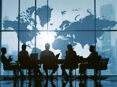 Das Bild zeigt eine eindrucksvolle Business-Szene im Breitbildformat mit starkem Hell-Dunkel-Kontrast (Silhouette): Im Vordergrund sitzen fünf Personen in Business-Kleidung an einem langen Konferenztisch, während sie auf Laptops und Mobilgeräten arbeiten oder beratschlagen. Der Raum wird von einer riesigen, bodentiefen Fensterscheibe dominiert, die den Blick auf eine städtische Skyline freigibt. Über die Glasfront ist in kühlen Blau- und Weißtönen eine große Weltkarte gelegt. Das Motiv vermittelt Themen wie globale Strategie, internationale Zusammenarbeit, Weltwirtschaft und Unternehmensführung.