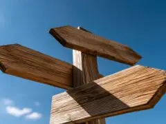 DaBild zeigt einen hölzernen Wegweiser mit drei blanko Pfeilschildern, die in unterschiedliche Richtungen weisen. Die Pfeile sind aus naturbelassenem, maserungsreichem Holz gefertigt und zeichnen sich vor einem strahlend blauen Himmel mit leichten Schönwetterwolken ab. Das Motiv symbolisiert Entscheidungsfindungen, Orientierung, Richtungswechsel oder strategische Optionen in Beruf und Leben.