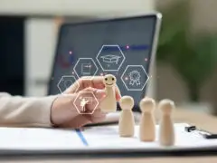 Das Bild zeigt ein Motiv zum Thema Personalentwicklung, Qualifizierung und Karriereförderung. Eine Hand greift nach einer lächelnden Holzspielfigur, die vor drei weiteren Holzfiguren auf Unterlagen auf einem Schreibtisch steht. Über der ausgewählten Figur sind grafische Hexagon-Icons schwebend eingeblendet, die Symbole für Bildung und Entwicklung zeigen (Glühbirne, Buch, Dozent, Doktorhut und Zertifikat/Ordensschleife). Im unscharfen Hintergrund sind ein Laptop und Zimmerpflanzen zu erkennen. Das Bild vermittelt anschaulich Themen wie gezielte Mitarbeiterförderung, Weiterbildung, Talentmanagement und Kompetenzaufbau.