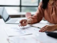 Das Bild zeigt eine fokussierte Arbeits- und Analyseszene im professionellen Büroumfeld. Im Zentrum stehen die Hände einer Frau in einer terracottafarbenen Bluse, die an einem hellen Schreibtisch sitzt. Sie hält einen Stift in der Hand und deutet damit auf ausgedruckte Berichte mit Diagrammen und Grafiken. Auf dem Tisch liegen zudem ein aufgeschlagenes Notizbuch, ein Taschenrechner sowie im unscharfen Hintergrund ein geöffneter Laptop und ein Kaffeebecher. Das Motiv vermittelt eine konzentrierte, strukturierte Arbeitsatmosphäre rund um die Themen Finanzplanung, Datenanalyse, Controlling und Strategieentwicklung.