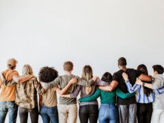 Das Bild zeigt eine Gruppe von neun Menschen, die nebeneinanderstehen und dem Betrachter komplett den Rücken zudrehen. Sie stehen eng beieinander vor einer schlichten, hellen Wand und legen jeweils die Arme um die Schultern oder den Rücken der neben ihnen stehenden Personen, was ein starkes Gefühl von Gemeinschaft, Zusammenhalt und Teamgeist vermittelt. Die Personen tragen alle lässige Alltagskleidung – wie Jeans, T-Shirts, Pullover oder Hemden – und sind in ihren Haarfarben, Frisuren und Hauttönen sehr unterschiedlich, was Vielfalt und Diversität ausstrahlt. Über den Köpfen der Gruppe bleibt die obere Hälfte des Bildes als schlichte, weiße Fläche frei.