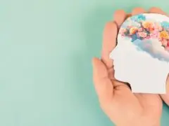 Das Bild zeigt eine ästhetische, symbolische Darstellung auf helltürkisem/pastellgrünem Hintergrund. Zwei Hände halten schützend einen Papierausschnitt in Form einer Kopf-Silhouette. Im oberen Bereich der Silhouette ist der Gehirnbereich mit einer farbenfrohen Illustration aus bunten Blumen, Blüten und Wolken gefüllt. Das Motiv steht sinnbildlich für Mental Health, emotionale Wohlbefinden, psychische Gesundheit, Achtsamkeit sowie kreatives und positives Denken im Berufs- und Lebenskontext.