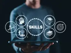 Das Bild zeigt ein Motiv zum Thema Kompetenzentwicklung, Qualifikation und berufliche Fähigkeiten. Eine Person hält flach ein Tablet in der Hand, über dem eine Reihe von digital leuchtenden, ineinandergreifenden Zahnrad-Icons schwebt. Im zentralen, größten Zahnrad steht das Wort „SKILLS“. Es ist umgeben von sechs kleineren Symbolen für Karriere/Aufstieg, Kopf mit Zahnrädern, Präsentation/Training, akademische Bildung (Doktorhut mit Glühbirne), Buch und Zielscheibe. Das Motiv symbolisiert Themen wie Skill-Management, kontinuierliches Lernen, Weiterbildung und Future Skills.
